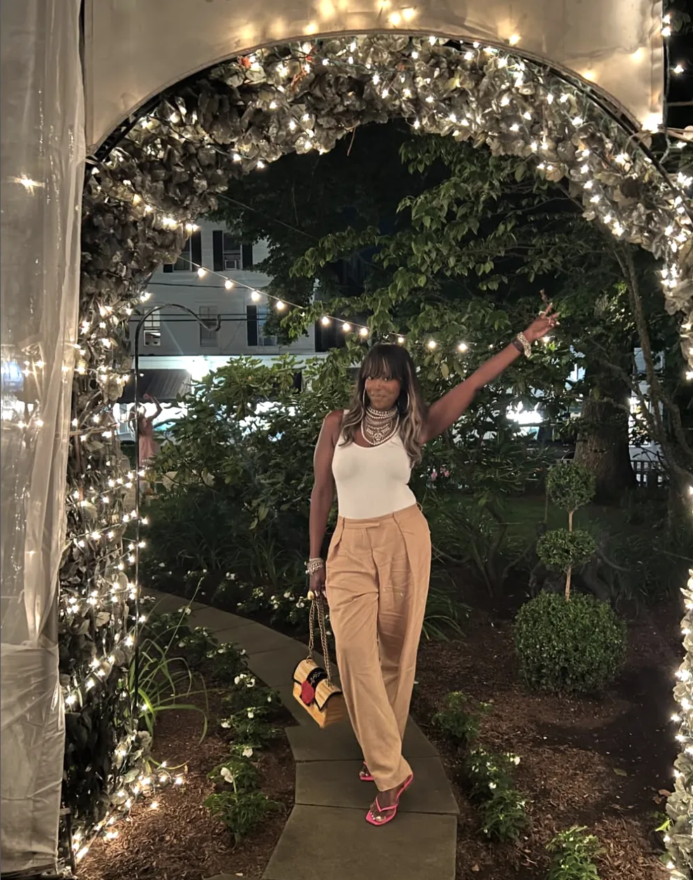 Mercedes under a lit garden arch of string lights and greenery at night, one arm raised, in a white tank, camel wide-leg trousers, pink heels and a small woven bag